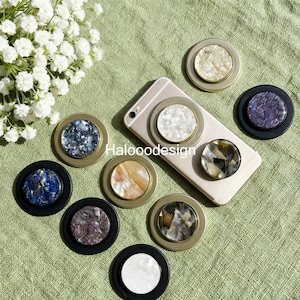 May include: A collection of decorative phone grips in various colors and designs, including white, blue, and iridescent patterns. The grips are round with a metallic gold or black border. The word "Haloooodesign" is visible on a phone.