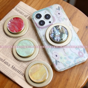 Op de afbeelding: Een verzameling telefoonhouders met een goudkleurige basis en kleurrijke, iriserende centra. Eén houder is bevestigd aan een telefoonhoesje met bloemenpatroon. De houders hebben rode, groene, gele en abalone schelp ontwerpen. De merknaam "Halooodesign" is zichtbaar.