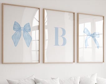 May include: Three framed wall art prints with light wood frames. The left print features a light blue bow, the center print displays a large light blue letter B, and the right print shows a smaller light blue bow on a white background.