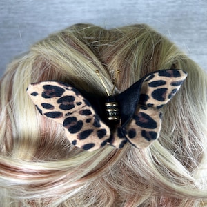 May include: A butterfly hair clip with a black body and tan wings patterned with brown spots. The clip has gold-colored antennae and a decorative gold-colored center. The hair accessory is attached to blonde hair.