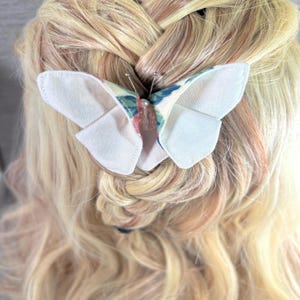 May include: A fabric butterfly hair clip with a floral pattern in shades of blue, green, and pink, attached to a blonde braid. The butterfly has white wings and a pearl-like center.
