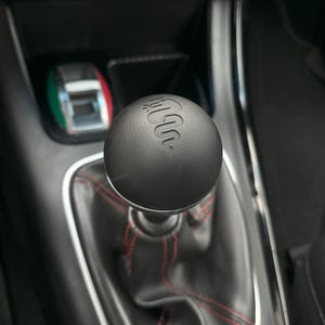 May include: A black leather gear shift knob with a red stitching detail and the Alfa Romeo logo embossed on the top.
