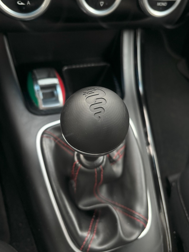 May include: A black leather gear shift knob with a red stitching detail and the Alfa Romeo logo embossed on the top.