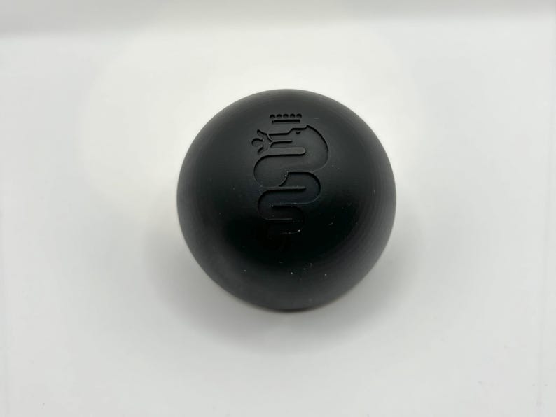 May include: A black round gear shift knob with a silver Alfa Romeo logo.