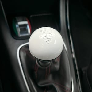 May include: A white gear shift knob with a silver Alfa Romeo logo. The knob is in a black car interior.