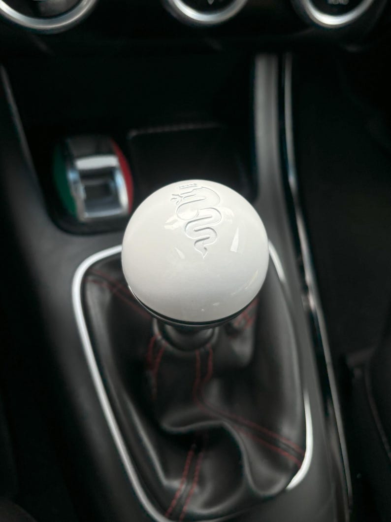 May include: A white gear shift knob with a silver Alfa Romeo logo. The knob is in a black car interior.