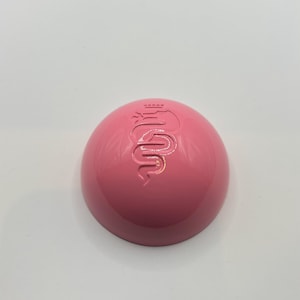 May include: A pink, round, plastic container with a raised, embossed logo of a snake with a crown on its head.
