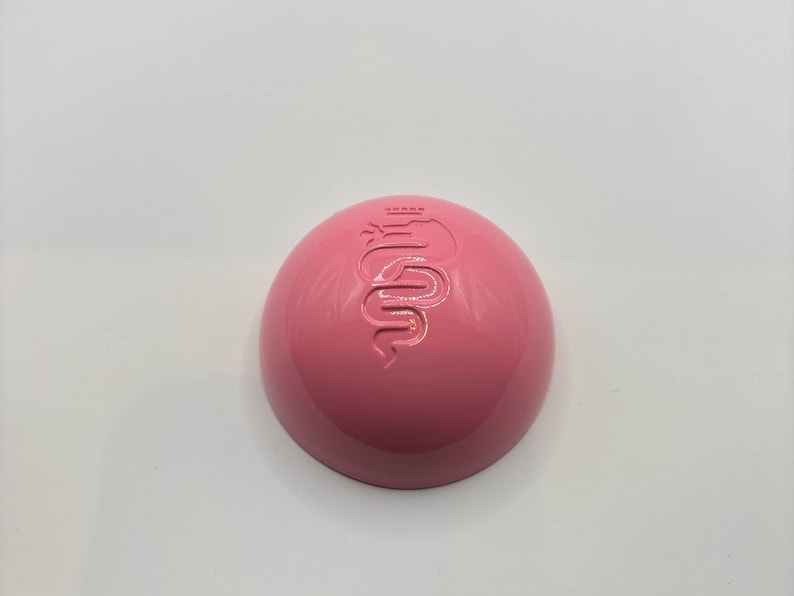 May include: A pink, round, plastic container with a raised, embossed logo of a snake with a crown on its head.