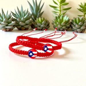 May include: Two red macrame bracelets with a Puerto Rican flag design. The bracelets are made of woven red cord and feature a round charm with the flag's emblem. The background includes green succulents.