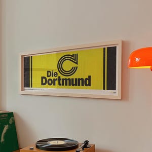 May include: A framed horizontal poster featuring a yellow background with black vertical stripes on the sides, a stylised black letter C logo, and the text Die Dortmund in bold black font, displayed on a wall above a wooden record player.