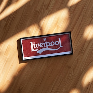 May include: A rectangular framed wall art piece featuring a red background with the white text Liverpool in a stylized script font. A small white emblem is positioned above the text. The frame is black and has a clean, simple border design.