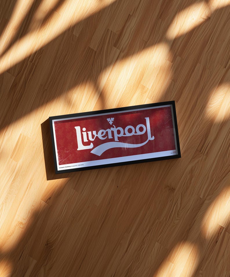 May include: A rectangular framed wall art piece featuring a red background with the white text Liverpool in a stylized script font. A small white emblem is positioned above the text. The frame is black and has a clean, simple border design.