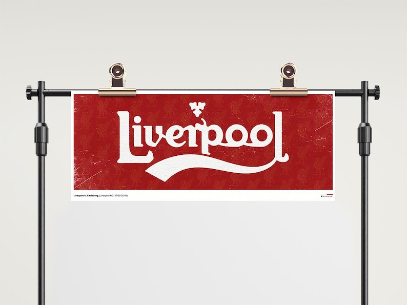 May include: A rectangular red banner featuring the white text Liverpool in a stylised font with a small white emblem above the letters. The background has a subtle repeating pattern and a distressed texture. The banner is held by two metal clips.
