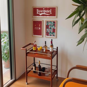 May include: A two-tiered wooden bar cart with metal railings and wheels, holding various glass bottles, two yellow lemons in a small bowl, and clear drinking glasses. The cart is positioned in front of a wall displaying three framed Liverpool posters.