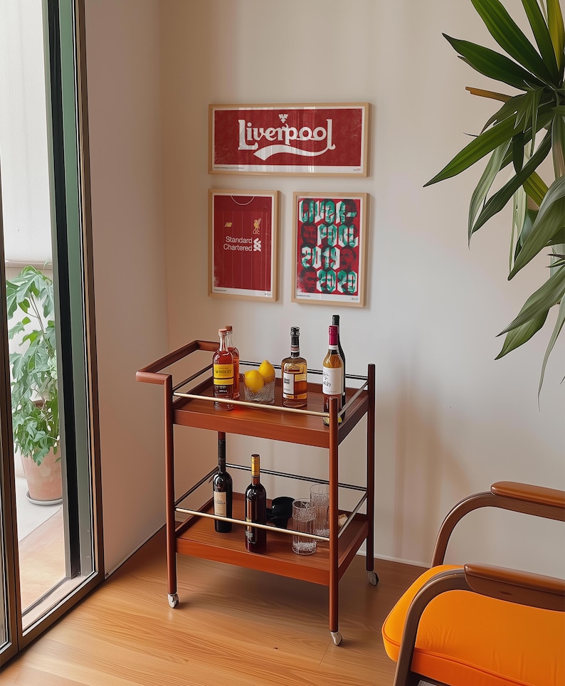 May include: A two-tiered wooden bar cart with metal railings and wheels, holding various glass bottles, two yellow lemons in a small bowl, and clear drinking glasses. The cart is positioned in front of a wall displaying three framed Liverpool posters.