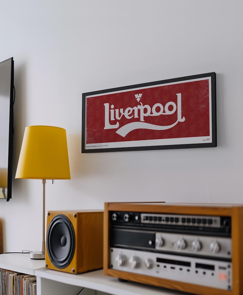 May include: A rectangular framed wall art piece featuring the word Liverpool in white stylized lettering against a textured red background. The design includes a small white emblem above the text and a white swoosh underline, all encased in a thin black frame.