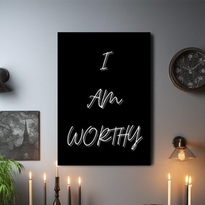 May include: A black and white motivational poster with the text "I am worthy" in a stylized font. The poster is hanging on a gray wall above a wooden table with candles and a black chair.