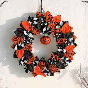 May include: A Halloween wreath made of fabric scraps in orange, black, and white. The wreath is decorated with a small orange jack-o'-lantern and black bats.