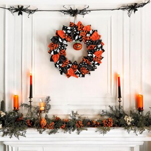 May include: A black, white, and orange Halloween wreath with a small pumpkin ornament hanging in the center. The wreath is decorated with black and white fabric bows and orange fabric. The wreath is hanging on a white wall above a fireplace mantel decorated with greenery and candles.