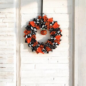 May include: A Halloween-themed wreath made of fabric scraps in black, orange, and white. The wreath features a variety of spooky designs, including bats, ghosts, and pumpkins. A small orange pumpkin ornament is hanging in the center of the wreath.