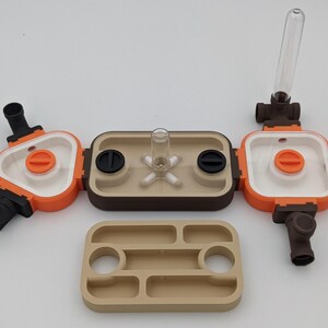 May include: A modular ant farm in orange, white, and brown, with a clear glass tube and a beige tray. The ant farm features multiple compartments and connecting tubes. The tray has recessed areas.