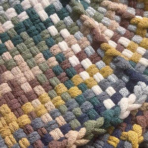 May include: Close-up of a woven textile with a basket weave pattern. The textile features a mix of colors, including shades of green, brown, yellow, and blue. The texture appears soft and the weave is tight, suggesting a handmade item.