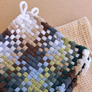 May include: A woven potholder in a square shape, featuring a mix of neutral colors including white, brown, beige, and green. The potholder has a loop for hanging and is placed on a yellow textured surface.