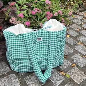 May include: A green and white gingham tote bag with white lining. The bag has two handles and a tag that reads "MADE BY MAISE". The bag is on a stone surface with pink flowers in the background.
