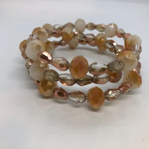 Könnte beinhalten: Ein dreireihiges Armband aus facettierten Glasperlen in den Farben Pfirsich, Creme und Klar. Die Perlen sind auf einem dünnen Draht aufgefädelt und haben einen zarten, femininen Look.