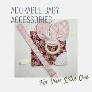 May include: Pink and white baby teether with a wooden ring and a floral fabric attached. The teether is attached to a pink and white strap with a clip.