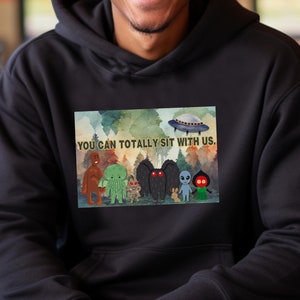 Könnte beinhalten: Ein schwarzer Hoodie mit einem grafischen Druck einer Gruppe von Cartoon-Monster und einem UFO. Der Text "You can totally sit with us." ist auf dem Hoodie gedruckt.