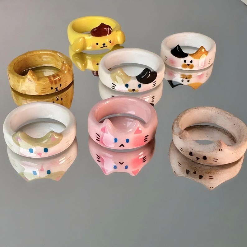 K&ouml;nnte beinhalten: Satz von zehn Keramikringen mit verschiedenen Katzendesigns. Die Ringe sind in verschiedenen Farben bemalt, darunter Gelb, Rosa, Wei&szlig; und Braun. Jeder Ring hat ein einzigartiges Katzengesichtsdesign mit verschiedenen Ausdr&uuml;cken.