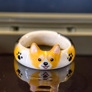May include: A white ceramic ring featuring a hand-painted Corgi face with orange and white fur, pointed ears, and a small pink tongue. The band is decorated with small black paw prints on either side of the dog's face, creating a detailed design.