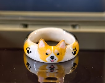 May include: A white ceramic ring featuring a hand-painted Corgi face with orange and white fur, pointed ears, and a small pink tongue. The band is decorated with small black paw prints on either side of the dog's face, creating a detailed design.