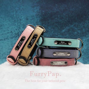 May include: Four personalised leather dog collars stacked on top of each other. The collars are in shades of pink, blue, grey, and light blue. Each collar has a silver name tag with a name and phone number.