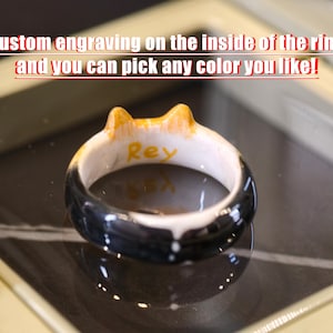 May include: A ceramic ring featuring a black, white, and orange cat-ear design with the name Rey engraved in yellow on the interior. The text Custom engraving on the inside of the ring and you can pick any color you like! is visible above.