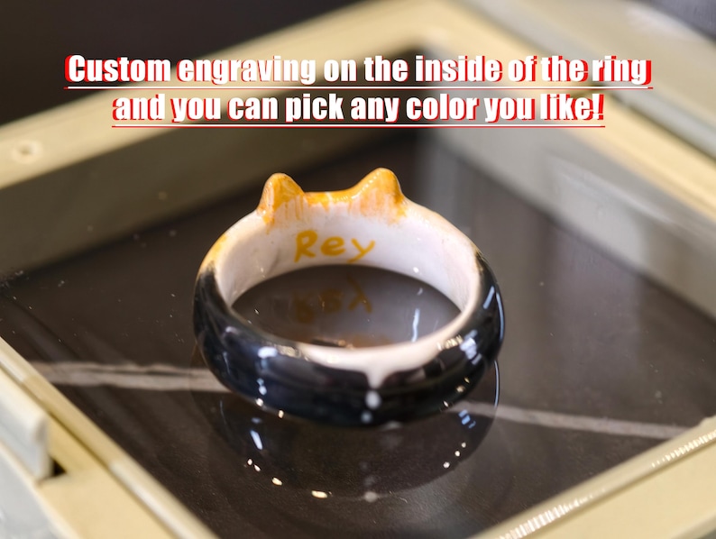 May include: A ceramic ring featuring a black, white, and orange cat-ear design with the name Rey engraved in yellow on the interior. The text Custom engraving on the inside of the ring and you can pick any color you like! is visible above.
