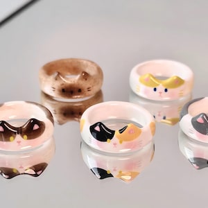 K&ouml;nnte beinhalten: F&uuml;nf Keramikringe mit verschiedenen Katzenk&ouml;pfen. Die Ringe sind wei&szlig;, braun und grau mit schwarzen, gelben und rosa Details.