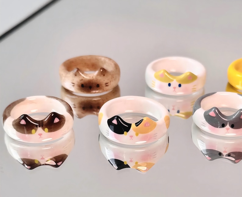 K&ouml;nnte beinhalten: F&uuml;nf Keramikringe mit verschiedenen Katzenk&ouml;pfen. Die Ringe sind wei&szlig;, braun und grau mit schwarzen, gelben und rosa Details.