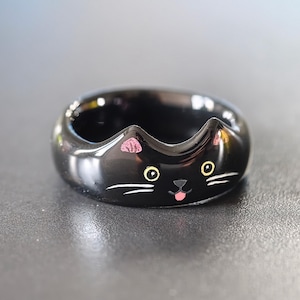 May include: A glossy black ring shaped like a cat face, featuring two pointed ears at the top, yellow eyes, a small black nose, a tiny pink tongue, and white whiskers painted on the front surface of the band.