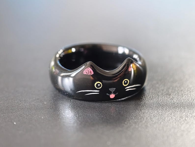May include: A glossy black ring shaped like a cat face, featuring two pointed ears at the top, yellow eyes, a small black nose, a tiny pink tongue, and white whiskers painted on the front surface of the band.