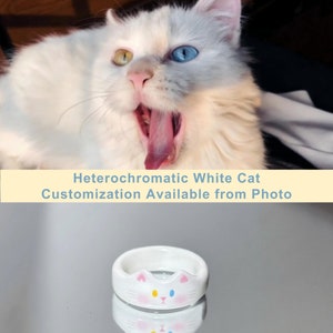 K&ouml;nnte beinhalten: Wei&szlig;er Keramikring mit einem Katzenkopfdesign. Die Katze hat ein blaues und ein gelbes Auge. Der Ring liegt auf einer reflektierenden Oberfl&auml;che.
