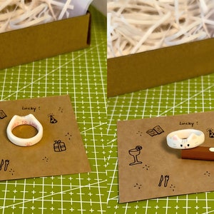 K&ouml;nnte beinhalten: Zwei wei&szlig;e Keramikringe mit unterschiedlichen Designs. Ein Ring tr&auml;gt die Aufschrift "WED TO EAT". Der andere Ring zeigt ein Katzen Gesicht mit rosa Ohren und einer rosa Nase. Beide Ringe liegen auf braunem Papier mit schwarzen Zeichnungen von Partyartikeln.