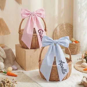 Puede incluir: Dos cestas de mimbre con grandes lazos a rayas. Uno es rosa con el nombre "Gisen" bordado, el otro azul claro con "Apollo". Las cestas son para Pascua o para regalar. Los lazos tienen un patrón de rayas verticales.