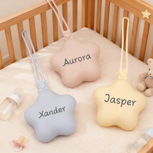 May include: Three star-shaped baby toys in pastel colors: pink, blue, and yellow. Each toy has a name printed on it: "Aurora", "Xander", and "Jasper". The toys have a loop for hanging and are placed on a crib sheet.