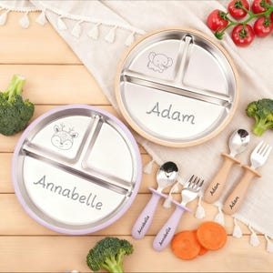 Peut inclure: Deux assiettes divisées en acier inoxydable, une avec un bord violet clair et le nom "Annabelle", l'autre avec un bord en bois et le nom "Adam". Des cuillères et des fourchettes assorties sont également incluses. Les assiettes ont des illustrations d'animaux.