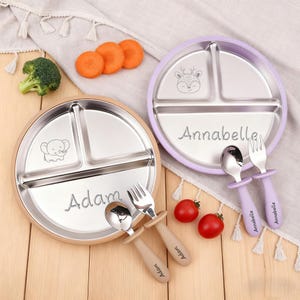 Peut inclure: Deux assiettes divisées en acier inoxydable avec des ustensiles assortis. L'une est dorée avec un éléphant et le nom "Adam". L'autre est violette avec un cerf et le nom "Annabella". Brocolis, carottes et tomates sont également présents.