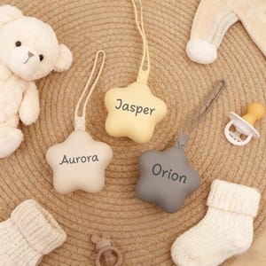 Op de afbeelding: Drie stervormige fopspeenclips voor baby's in crème, geel en grijs, elk met een naam erop gedrukt: Aurora, Jasper en Orion. De clips hebben een lus om aan een fopspeen te bevestigen. Andere babyartikelen staan op de achtergrond.
