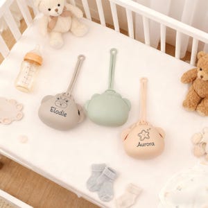 May include: Three baby pacifier holders in neutral colors: taupe, sage green, and peach. Each holder has a bear-shaped design with a loop for attachment. The taupe holder says "Elodie", and the peach holder says "Aurora".