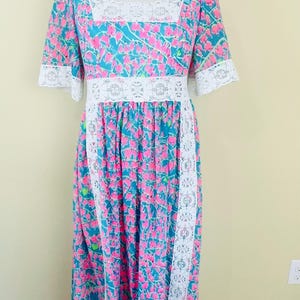 Pu&ograve; includere: Un abito lungo fino ai piedi con sfondo color ottanio e un motivo floreale rosa ripetuto. Il capo presenta bordi in pizzo bianco lungo lo scollo quadrato, i polsini delle maniche, il punto vita e i pannelli verticali della gonna.